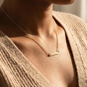 May include: A gold-colored bar necklace with a delicate chain. The rectangular pendant has the word "VIVIII" engraved on it. The necklace is worn on a person wearing a beige sweater.