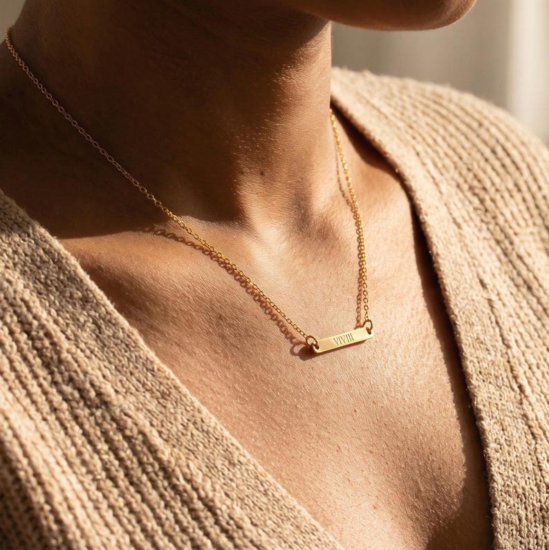 May include: A gold-colored bar necklace with a delicate chain. The rectangular pendant has the word "VIVIII" engraved on it. The necklace is worn on a person wearing a beige sweater.