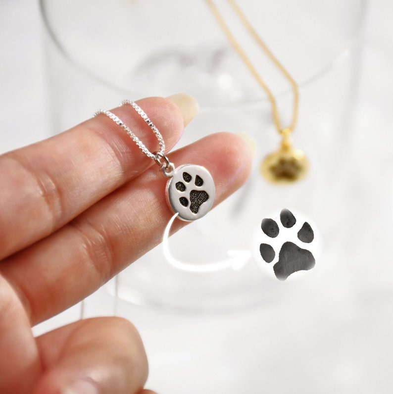 Op de afbeelding: Een zilveren ketting met een ronde hanger met een pootafdrukontwerp. De hanger wordt door een hand vastgehouden, met een gouden ketting op de achtergrond. Een pootafdruk-afbeelding wordt ook getoond.