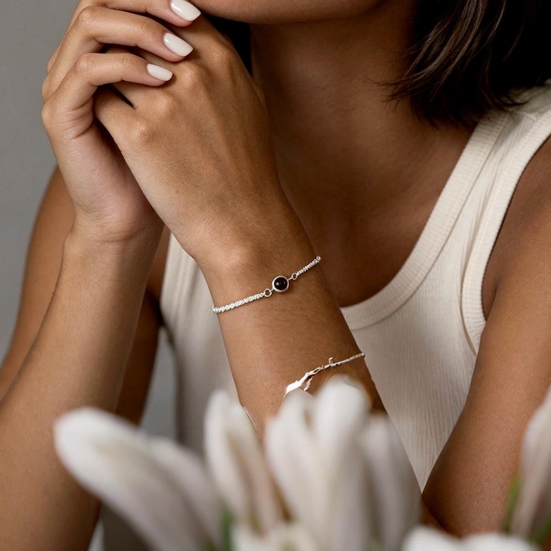 May include: A silver bracelet with a small, round, dark gemstone is worn on a wrist. The bracelet has a delicate chain design. Another silver bracelet is also visible.