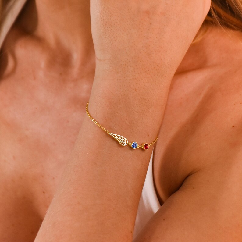 Pode incluir: Uma delicada pulseira de ouro com um berloque de asa e dois detalhes de pedras preciosas. A pulseira tem uma pequena pedra preciosa azul e vermelha. A pulseira &eacute; usada no pulso.