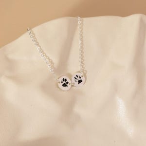May include: A silver necklace with two circular pendants, each showing a black paw print. The pendants are linked by a fine chain, creating a simple yet meaningful accessory. The necklace is displayed on a cream-coloured surface.
