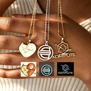May include: Three necklaces with pendants in gold and silver tones. One pendant is a heart shape with two cat figures, another is a circle with a stylized "S", and the third is a geometric design with the word "KOMPUTE".