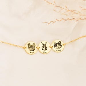 May include: A gold necklace with three circular pendants, each engraved with a pet portrait: a dog, a rabbit, and a cat. The names 'Hugo', 'Diesl', and 'Sunny' are below each image. The necklace is displayed on a light background.