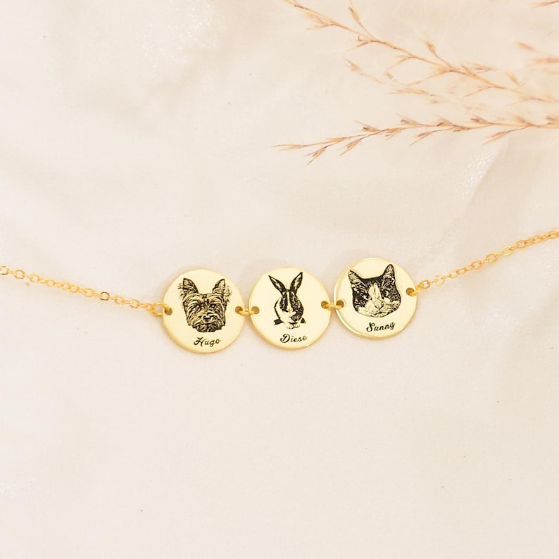May include: A gold necklace with three circular pendants, each engraved with a pet portrait: a dog, a rabbit, and a cat. The names 'Hugo', 'Diesl', and 'Sunny' are below each image. The necklace is displayed on a light background.