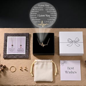 May include: An assortment of jewelry and gift boxes arranged on a textured surface. The items include a gold necklace in a black box, a white gift box with a bow design, a small ring, earrings, and a drawstring bag. A card reads "Best Wishes".