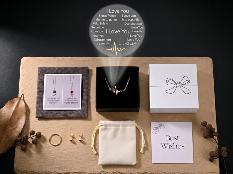 May include: An assortment of jewelry and gift boxes arranged on a textured surface. The items include a gold necklace in a black box, a white gift box with a bow design, a small ring, earrings, and a drawstring bag. A card reads "Best Wishes".