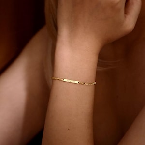 May include: A gold-toned chain bracelet featuring a thin, rectangular horizontal bar plate. The center of the bar is engraved with the Roman numerals VII XXIII. The bracelet is constructed with a delicate link chain that secures around the wrist.