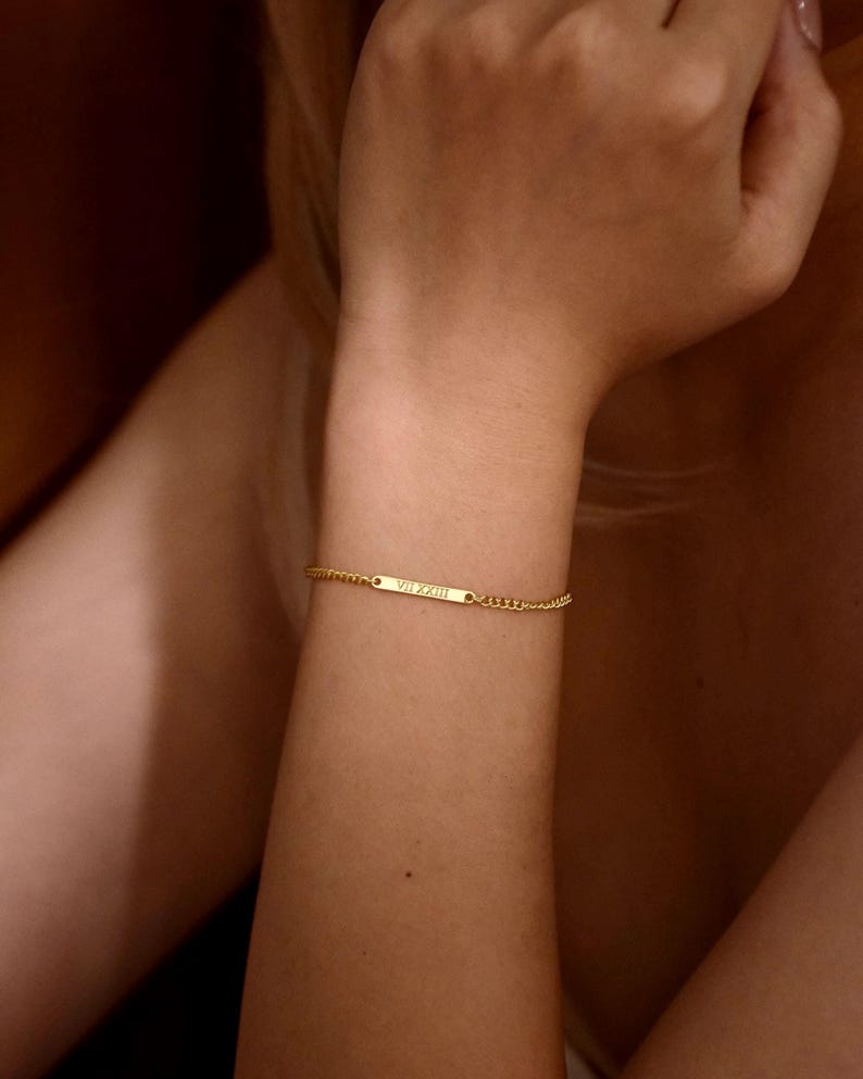May include: A gold-toned chain bracelet featuring a thin, rectangular horizontal bar plate. The center of the bar is engraved with the Roman numerals VII XXIII. The bracelet is constructed with a delicate link chain that secures around the wrist.