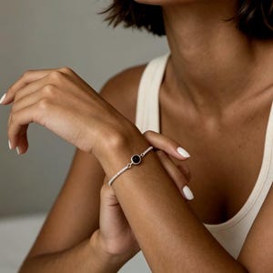 May include: A silver bracelet with a black circular stone in the center. The bracelet is adorned with small, sparkling stones. The jewelry is worn on a wrist.