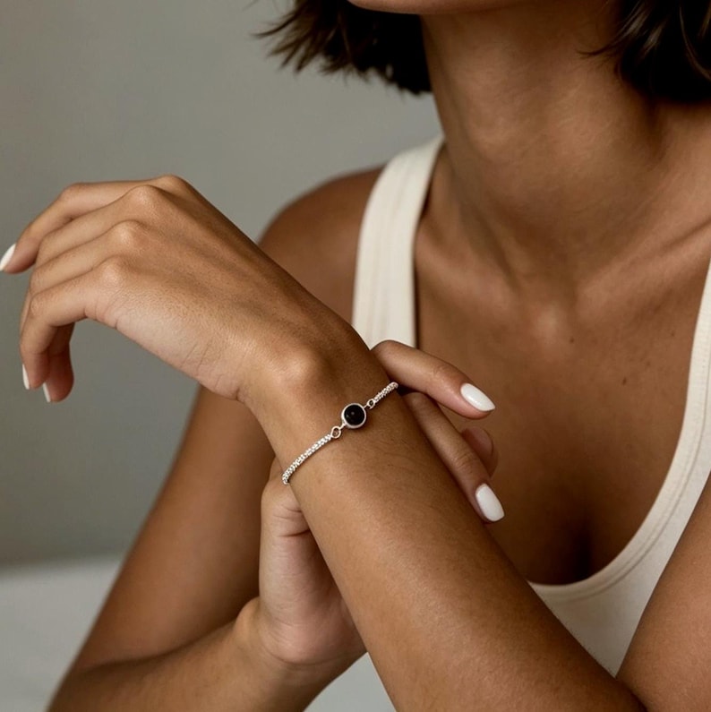 May include: A silver bracelet with a black circular stone in the center. The bracelet is adorned with small, sparkling stones. The jewelry is worn on a wrist.