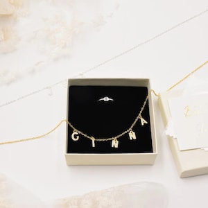 May include: A white box with a black interior containing a gold necklace with the letters "GINNA" hanging from it. A silver ring with a single diamond is also in the box. The box is sitting on a white surface.