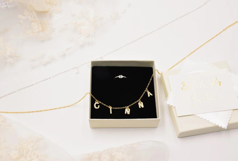 May include: A white box with a black interior containing a gold necklace with the letters "GINNA" hanging from it. A silver ring with a single diamond is also in the box. The box is sitting on a white surface.