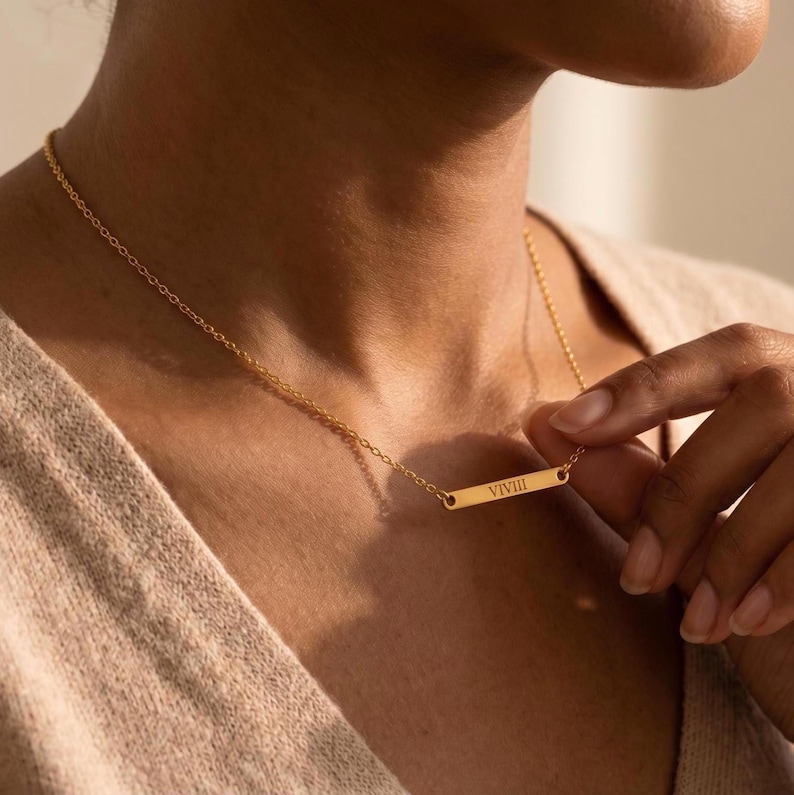 May include: A gold-colored bar necklace with the word "VIIII" engraved on it. The necklace is on a delicate chain. The person is wearing a beige sweater.