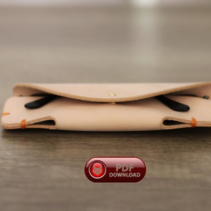 Puede incluir: Un estuche de gafas de cuero de color beige claro con unas gafas negras en su interior. El estuche se cierra con un broche y tiene costuras naranjas. El estuche est&aacute; sobre una superficie de madera.