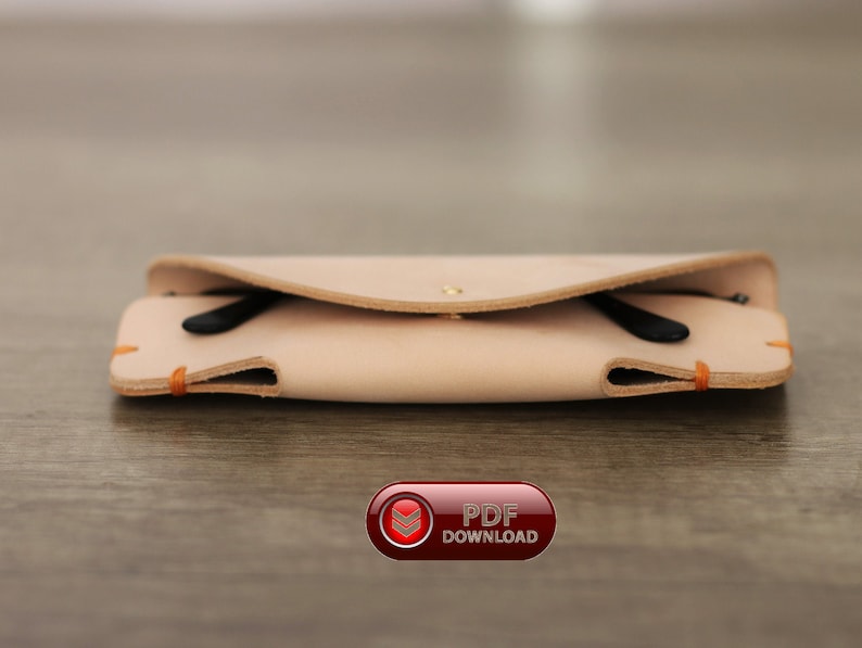Puede incluir: Un estuche de gafas de cuero de color beige claro con unas gafas negras en su interior. El estuche se cierra con un broche y tiene costuras naranjas. El estuche est&aacute; sobre una superficie de madera.