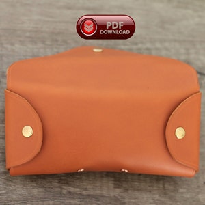 Puede incluir: Estuche de gafas de cuero marr&oacute;n anaranjado abierto, con cierres de bot&oacute;n a presi&oacute;n dorados. El estuche rectangular tiene bordes redondeados y una solapa curva. Un icono de descarga PDF rojo y blanco est&aacute; visible.