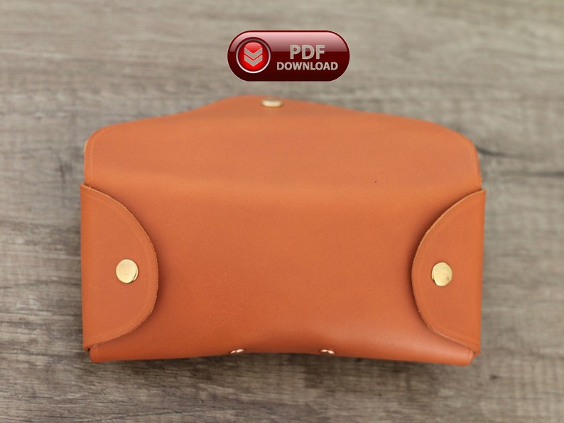 Puede incluir: Estuche de gafas de cuero marr&oacute;n anaranjado abierto, con cierres de bot&oacute;n a presi&oacute;n dorados. El estuche rectangular tiene bordes redondeados y una solapa curva. Un icono de descarga PDF rojo y blanco est&aacute; visible.
