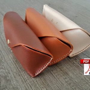 May include: Three leather eyeglass cases in shades of brown and cream. The cases are closed, with a snap closure on the darkest brown case. The lightest case has the initials "DK" embossed on it. The cases are on a gray wooden surface.