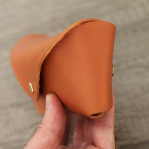 Puede incluir: Un estuche de gafas de cuero color canela. El estuche es curvo y tiene una solapa que se pliega sobre la parte superior. Dos peque&ntilde;os remaches dorados aseguran el estuche. El estuche se sostiene en una mano.