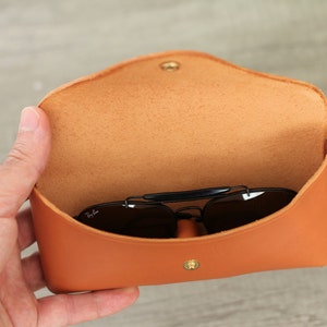 Puede incluir: Estuche de gafas de sol de cuero naranja abierto con cierre de bot&oacute;n a presi&oacute;n dorado. El interior es de color marr&oacute;n claro. Las gafas de sol negras son visibles dentro del estuche.