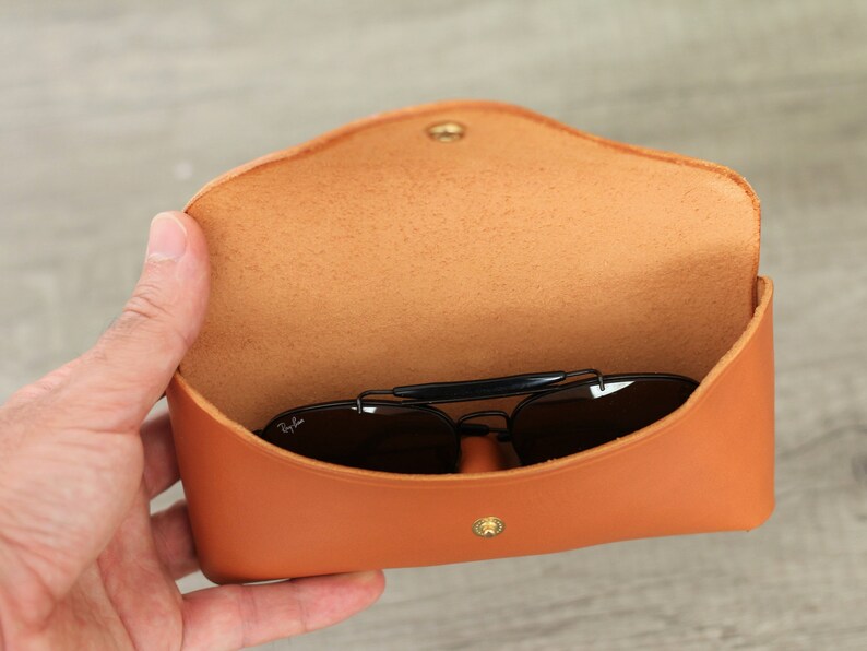 Puede incluir: Estuche de gafas de sol de cuero naranja abierto con cierre de bot&oacute;n a presi&oacute;n dorado. El interior es de color marr&oacute;n claro. Las gafas de sol negras son visibles dentro del estuche.