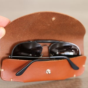 Puede incluir: Gafas de sol aviador negras dentro de un estuche de cuero marr&oacute;n. El estuche es de color marr&oacute;n medio c&aacute;lido con cierre de bot&oacute;n a presi&oacute;n. Las gafas tienen montura plateada y lentes oscuras.