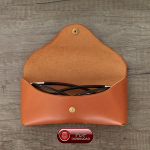 Puede incluir: Estuche de gafas de cuero marr&oacute;n claro abierto, con cierre de bot&oacute;n a presi&oacute;n. El estuche contiene un par de gafas con montura negra. El estuche est&aacute; sobre una superficie de madera.