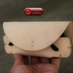 Puede incluir: Un estuche de gafas de cuero de color beige claro con una solapa curva y un cierre de bot&oacute;n dorado. El estuche tiene detalles de costuras naranjas y contiene unas gafas de sol negras. Tambi&eacute;n se ve un bot&oacute;n rojo con el texto "PDF DOWNLOAD".