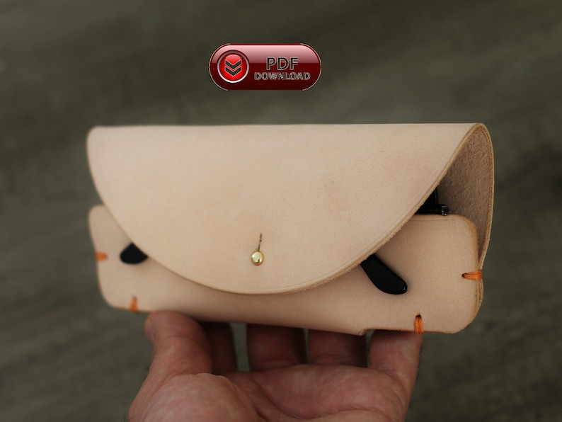 Puede incluir: Un estuche de gafas de cuero de color beige claro con una solapa curva y un cierre de bot&oacute;n dorado. El estuche tiene detalles de costuras naranjas y contiene unas gafas de sol negras. Tambi&eacute;n se ve un bot&oacute;n rojo con el texto "PDF DOWNLOAD".