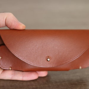Puede incluir: Un estuche de gafas de cuero marr&oacute;n con cierre de solapa curvada y un peque&ntilde;o broche met&aacute;lico. El estuche rectangular tiene bordes redondeados y costuras visibles. El estuche se sostiene en una mano.