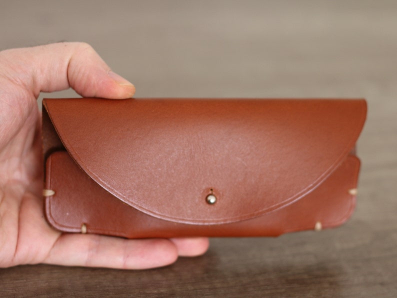 Puede incluir: Un estuche de gafas de cuero marr&oacute;n con cierre de solapa curvada y un peque&ntilde;o broche met&aacute;lico. El estuche rectangular tiene bordes redondeados y costuras visibles. El estuche se sostiene en una mano.