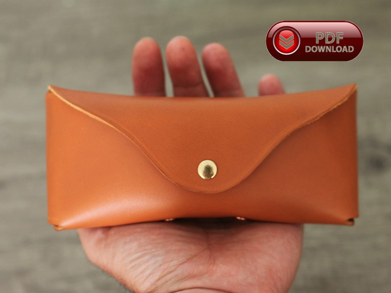 Puede incluir: Un estuche rectangular de cuero marr&oacute;n anaranjado, con una solapa curva y un cierre de bot&oacute;n a presi&oacute;n dorado. El estuche se sostiene en una mano y tiene una apariencia suave y pulida. La imagen tambi&eacute;n incluye un bot&oacute;n "PDF DOWNLOAD".