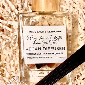 May include: A clear glass bottle with a wooden lid and a white label that reads "MINDTALITY SKINCARE I Can Love Me Better Than You Can VEGAN DIFFUSER WITH ROSE & STRAWBERRY QUARTZ HANDMADE IN AUSTRALIA 100ML". The bottle is filled with a clear liquid and contains a small piece of pink and red crystal. A black diffuser stick is lying on top of the bottle.