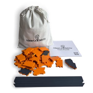 May include: A team puzzle set with orange and gray puzzle pieces, a drawstring bag, and a booklet. The bag and booklet feature the text "ideen.k³llektiv" and a lightbulb logo. The puzzle pieces are scattered around the bag.