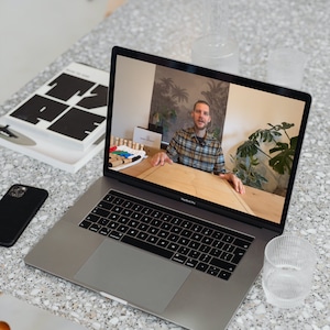 Puede incluir: Una persona en una videollamada en un ordenador portátil plateado. La persona lleva una camisa de cuadros y está sentada en una mesa de madera. La pantalla del ordenador portátil muestra una mesa de madera con una caja y algunos objetos coloridos sobre ella.