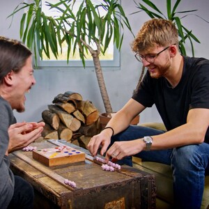 May include: Two people play a board game with pink plastic pieces on a wooden board. The board has a grid of colored circles and a small wooden box with a lid. The game is set on a wooden table with a pile of logs in the background.