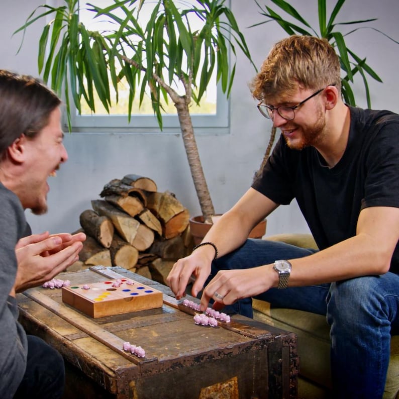 May include: Two people play a board game with pink plastic pieces on a wooden board. The board has a grid of colored circles and a small wooden box with a lid. The game is set on a wooden table with a pile of logs in the background.