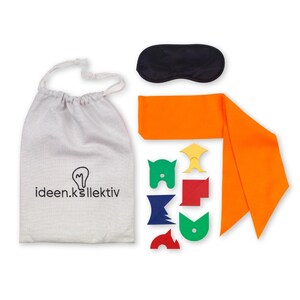 Può includere: Un set di pezzi di puzzle colorati in una borsa a coulisse bianca con il testo "ideen.kollektiv" su di essa. Il set include anche una maschera per dormire nera e una sciarpa di stoffa arancione.