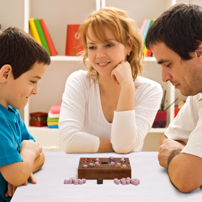 May include: A wooden game board with a hole in the center and colorful marbles. The board is surrounded by a group of people, including a child, a woman, and a man.