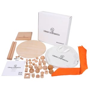 Könnte beinhalten: Ein Holzspielzeug-Set mit verschiedenen geometrischen Formen, einer runden Holzplattform, zwei Holzdübeln und einem orangefarbenen Stoff. Das Set enthält eine weiße Box und eine weiße Tasche, beide mit dem Text "ideen.k.llektiv".