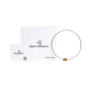 Op de afbeelding: Een zilverkleurige Team Ring ketting met drie goudkleurige kralen. De ketting wordt gepresenteerd met een witte doos en kaart, beide met het "ideen.k" logo en de woorden "Team Ring."