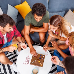 May include: A group of people playing a wooden board game with colorful marbles. The game board has a grid pattern and is surrounded by pink and white marbles.