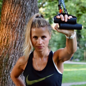 Pu&ograve; includere: Una donna indossa una canotta nera con un logo Nike dorato a forma di swoosh e tiene in mano una fascia di allenamento in sospensione nera e arancione. &Egrave; in piedi davanti a un albero.