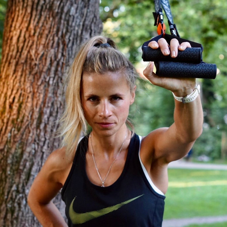 Pu&ograve; includere: Una donna indossa una canotta nera con un logo Nike dorato a forma di swoosh e tiene in mano una fascia di allenamento in sospensione nera e arancione. &Egrave; in piedi davanti a un albero.