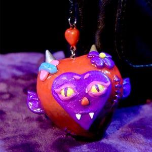 May include: A heart-shaped charm with a devilish design. The charm is red with a purple heart-shaped face, yellow eyes, and small white fangs. It features small horns, flowers, and a dangling red heart. Attached to a silver chain. A unique accessory.