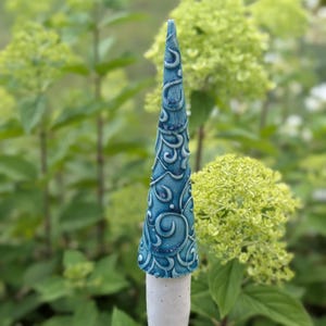 Könnte beinhalten: Ein konischer Gartenstecker aus Keramik mit einer glänzenden blauen Glasur und erhabenen, wirbelnden Reliefmustern. Der obere Teil ist ein spitzer blauer Kegel, während die Basis ein massiver, mattweißer zylindrischer Verbinder auf einem Holzstab ist.