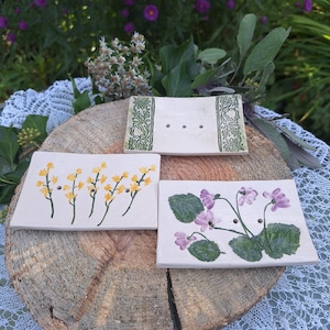 Könnte beinhalten: Drei handgefertigte Seifenschalen aus Keramik. Eine hat ein grünes Blumendesign, eine weitere gelbe Blumen und die dritte lila Blumen. Jede Schale hat drei Abflusslöcher und steht auf einer Holzoberfläche.