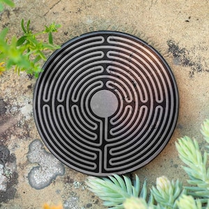 Puede incluir: Un posavasos circular negro con un diseño de laberinto plateado. El posavasos está sobre una superficie marrón claro con plantas verdes.