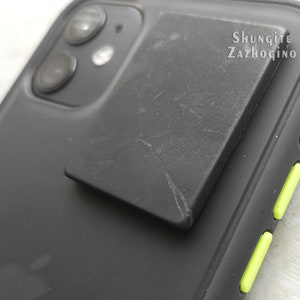 May include: A black phone case with a black square shungite stone attached to the back. The text "Shungite Zazhogino" is printed on the stone.